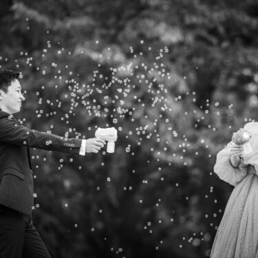 木々の前でシャボン玉を飛ばすバブルガンを向け合う新郎新婦のモノクロ写真。笑顔あふれる洋装前撮り