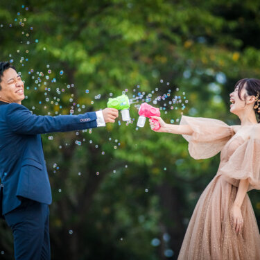 緑の木々を背景に、カラフルなバブルガンでシャボン玉を撃ち合うタキシードの新郎とカラードレスの花嫁の前撮り