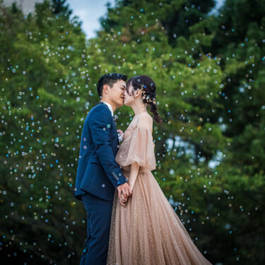 シャボン玉が舞う公園の木々の前でキスを交わすタキシードの新郎とカラードレスの花嫁のフォトウェディング
