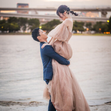 お台場の水辺で花嫁を高く抱き上げ、見上げて笑い合う新郎新婦。夕暮れの洋装ロケーションフォト