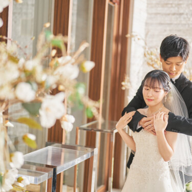ドライフラワーの装花越しに、花嫁を後ろから包み込むように抱きしめるタキシードの新郎。チャペルでのバックハグの結婚写真