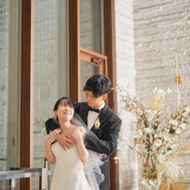 窓辺に立ち、後ろから花嫁を抱きしめる新郎と、その腕に手を添えて笑う花嫁。チャペルで撮るバックハグの前撮りポーズ