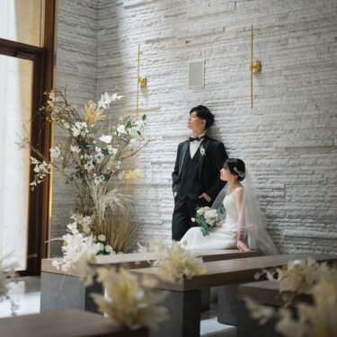 窓から光が差し込むチャペルの壁際で、壁にもたれる新郎とベンチに座るベールの花嫁。ドライフラワーの装花が彩る結婚写真