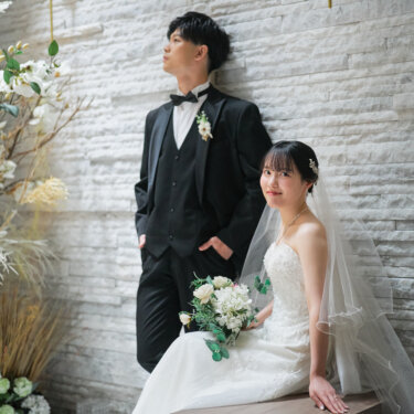 石積みの壁にもたれて遠くを見る新郎と、ベンチに座りブーケを持って振り向く花嫁。チャペルで撮るおしゃれな前撮りポーズ