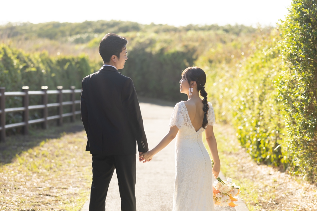 城ヶ島の緑の小道を手をつないで歩く、背中の開いたウェディングドレスの花嫁と新郎の後ろ姿