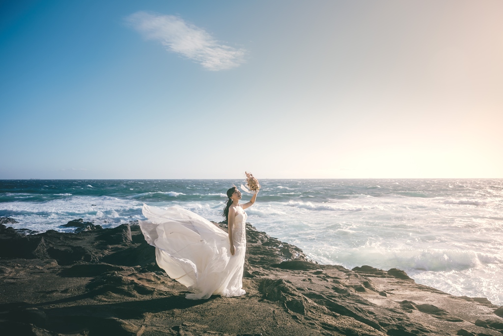 城ヶ島の岩場で海風にウェディングドレスとベールがなびく花嫁と新郎。海と空を背景にした洋装前撮り