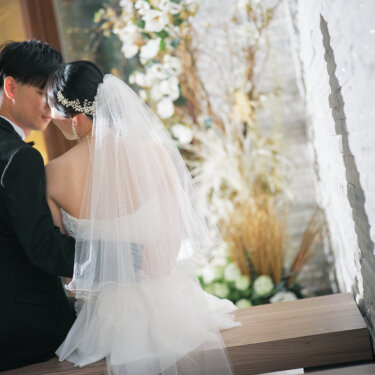 ベンチに並んで座り、ベール越しに顔を寄せ合う新郎新婦の後ろ姿。ドライフラワーの装花と石壁のチャペルで撮るフォトウェディング