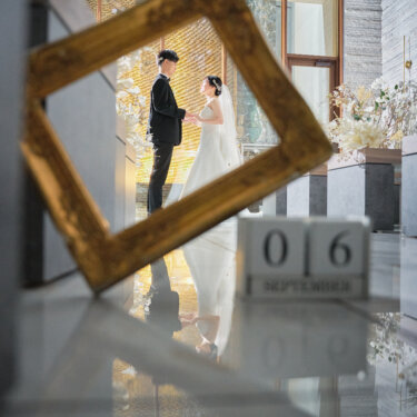 手前に置いた額縁と日付ブロックの奥に、向かい合う新郎新婦が小さく写るチャペルの前撮り。床に映り込むリフレクションもおしゃれな構図