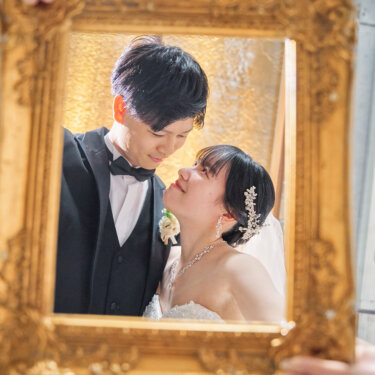 アンティークのゴールドの額縁越しに、見つめ合って微笑むタキシードの新郎とビジューのドレスの花嫁。額縁を使ったおしゃれなチャペルフォト