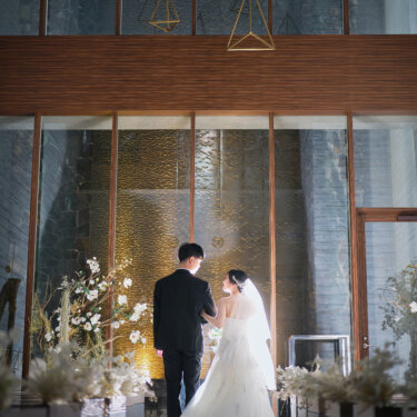 磨かれた床にドレスが映り込むバージンロードで、祭壇へ向かって並ぶ新郎新婦の後ろ姿。吹き抜けのチャペルで撮るフォトウェディング
