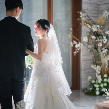 窓辺の逆光に照らされ、新郎の腕に手を添えて見上げるベール姿の花嫁。装花に囲まれたチャペルで撮るロマンチックな結婚写真