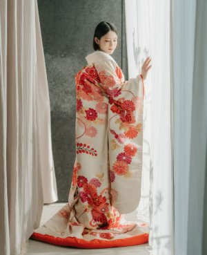 白地に赤い牡丹や桜を散らした色打掛で窓辺に立つ、表参道店の和装前撮りの花嫁（衣装番号DS01）