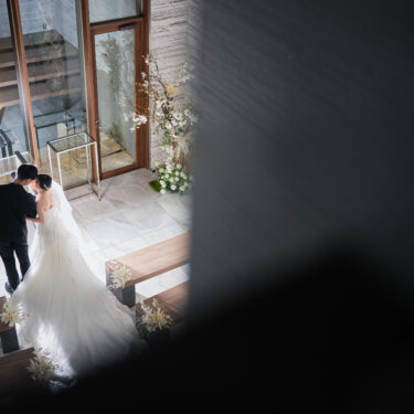 チャペルのガラス扉の前で寄り添う新郎新婦を、上階から壁越しにのぞき込むように撮った映画風のウェディングフォト
