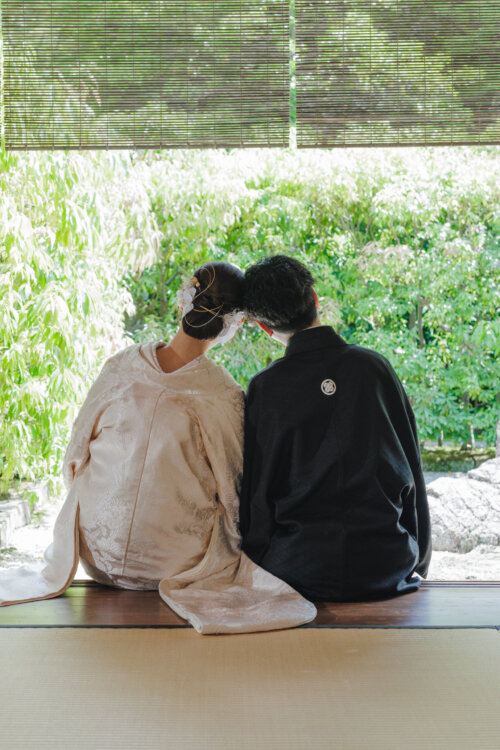 すだれ越しに緑の庭を眺め、縁側に並んで座る白無垢の花嫁と紋付の新郎の後ろ姿。和装前撮りの人気ポーズ