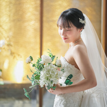 ロングベールにビジューの髪飾りをつけ、白いブーケを見つめて微笑む花嫁。金色の光が差すチャペルで撮る花嫁ソロの前撮り