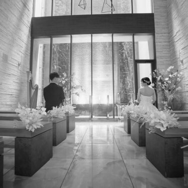 ガラスのオブジェが吊るされたチャペルの祭壇前に座る新郎新婦を、ベンチ越しに撮ったモノクロのウェディングフォト