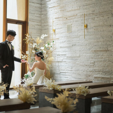 窓辺の光が差し込むチャペルで、ベンチに座る花嫁の手を取る新郎。ドライフラワーの装花に囲まれたロマンチックな前撮りポーズ