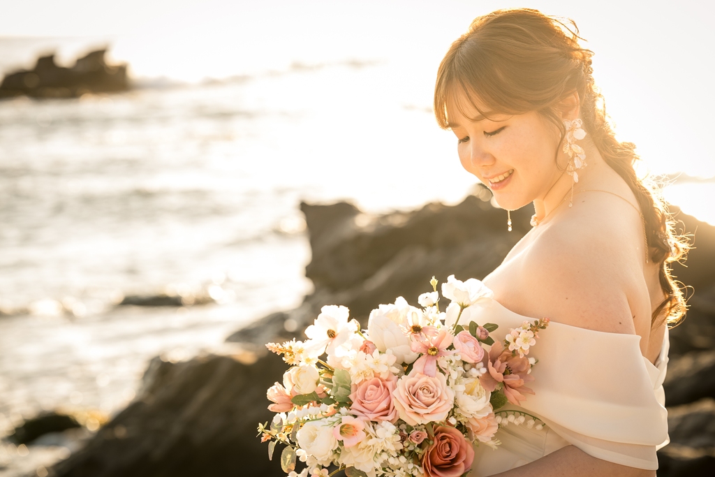 夕日の光の中でブーケを持ってほほえむ、オフショルダーのウェディングドレスの花嫁のアップ。城ヶ島での前撮り