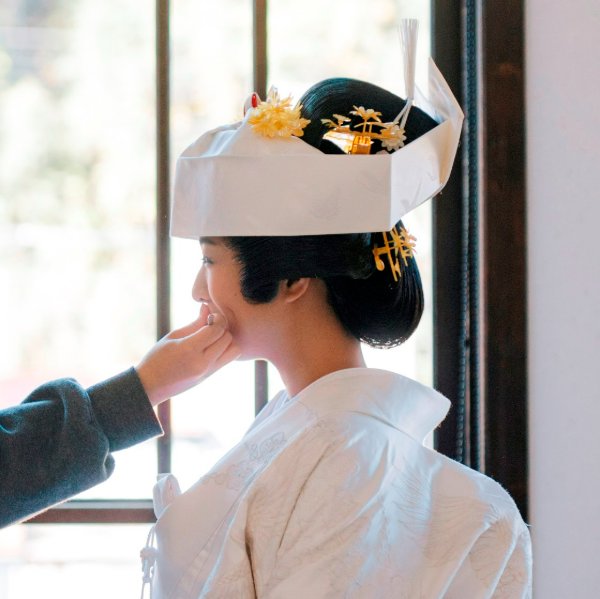 綿帽子と金の花かんざしを着けた白無垢姿の花嫁の横顔、リップメイクの最終仕上げをするスタッフの手