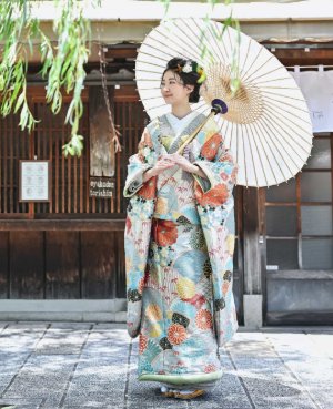 淡いブルーに草花を描いた懐かしい色合いの色打掛「山流水色」で和傘を差し、柳の揺れる街並みに立つ花嫁の和装前撮り（衣装番号SMOS11）