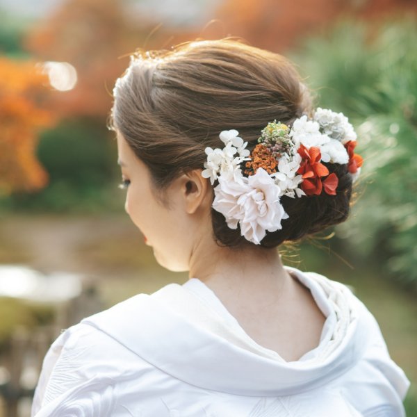 紅葉を背景に赤と白の花の髪飾りを添えた低めのシニヨン。白無垢の襟元に映える和装前撮りのヘアアレンジ