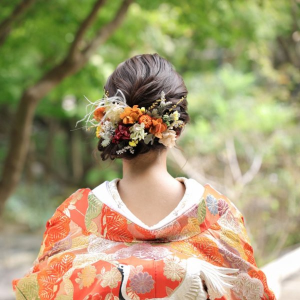 緑の木々を背景に、ドライフラワーの髪飾りをまとめ髪に添えた赤い色打掛の花嫁の後ろ姿。和装前撮りのヘアアレンジ