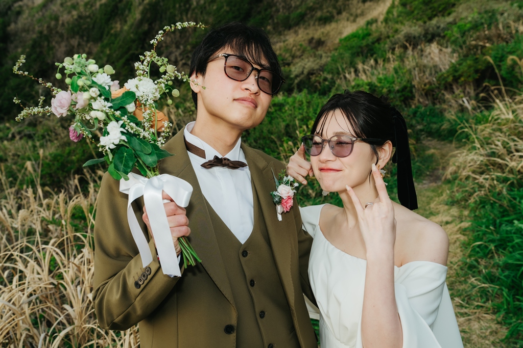 城ヶ島の草原で、グリーンと花のナチュラルなブーケを持ちサングラスをかけて笑う新郎新婦