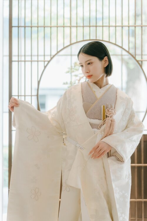 丸窓と格子戸のある和室で袖を広げ、桜の刺繍を見せる白無垢の花嫁。柔らかな光が差す和装前撮りの白無垢AS102