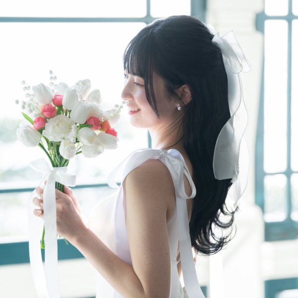 白いリボンを結んだ黒髪のダウンスタイルで、生花のブーケを持つウェディングドレスの花嫁の横顔。ナチュラルな前撮りヘア
