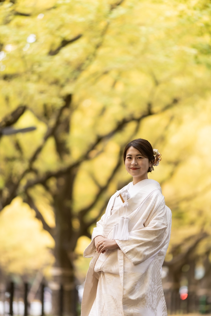 外苑いちょう並木の黄色い木々を背景に立つ、洋髪に花の髪飾りを合わせた白無垢の花嫁の全身写真