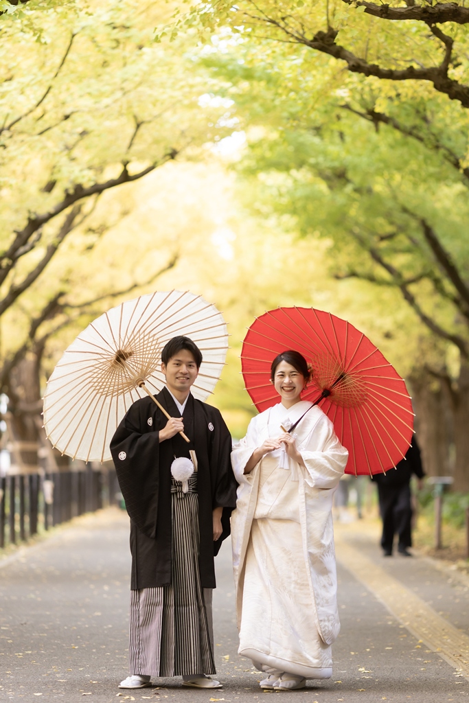 外苑いちょう並木で白と赤の和傘を差して並ぶ白無垢の花嫁と紋付袴の新郎。和傘を使った秋の和装コーディネート