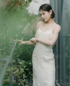 エリザベスフィルモアの手刺繍が繊細なスレンダーラインのウェディングドレスで、窓辺の緑の前で手を伸ばす表参道店の前撮り（衣装番号DE03）