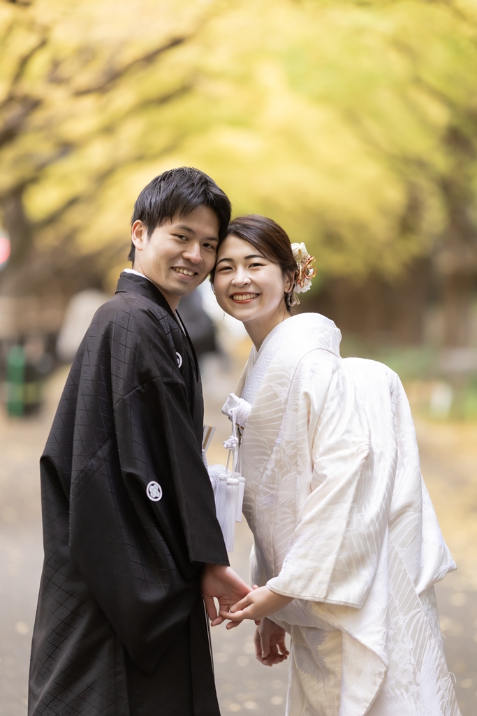 外苑いちょう並木の木の下で顔を見合わせて笑い合う白無垢の花嫁と紋付袴の新郎