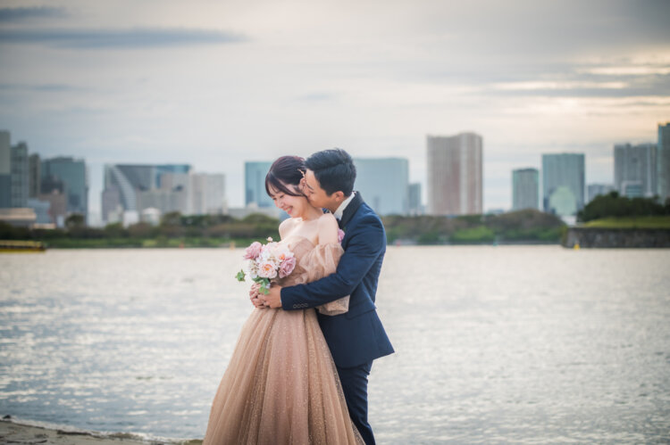 お台場海浜公園の水辺で、ベージュのチュールのカラードレスの花嫁を後ろから抱きしめる新郎。ビル群を望む洋装前撮り