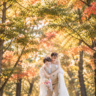 赤や黄色に色づいた並木の下で、ウェディングドレスの花嫁をおんぶして笑い合うベージュのベスト姿の新郎。秋の洋装前撮り