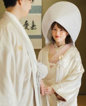 掛け軸のある和室で綿帽子をかぶり、白紋付の新郎と手を取って微笑む白無垢の花嫁。和装前撮り（白無垢／衣装番号SF105）